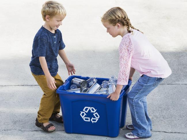 A young girl and her little brother standing on the driveway with a blue recycling bin full of empty bottles and cans. The bin carries the well-known ‘chasing arrows’ recycling logo. 