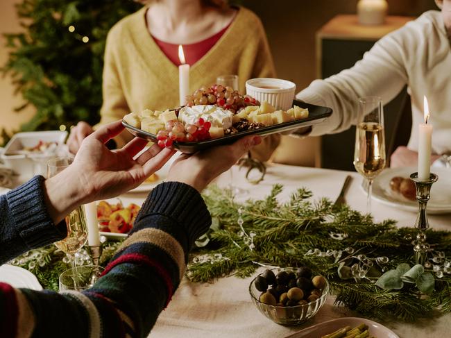 Medium close up of unrecognizable friend offering cheese plate to his friend while they sitting at festive table with pine garland on top