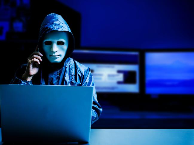 Anonymous computer hacker in white mask and hoodie. Obscured dark face using laptop computer for cyber attack and calling on cellphone, Data thief, internet attack, darknet and cyber security concept.