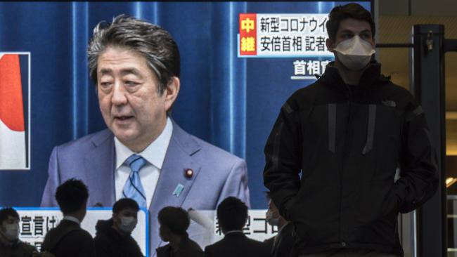 Pedestrians wearing face masks walk past a monitor showing a press conference by Japanese Prime Minister Shinzo Abe on Covid-19. Picture: Getty