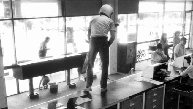 Still of security footage from an armed hold up at the NAB in Wynnum, September 1987. A man wearing a helmet and brandishing a pump-action shotgun jumps on the counter and demands money while a young boy cowers in fear under a table and customers freeze in their tracks.