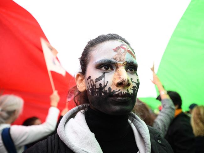 TOPSHOT - A protester wearing face-paint depicting France's iconic "Marianne" leading an uprising, stands under a giant Iranian flag as people gather in support of Kurdish woman Mahsa Amini during a protest on October 2, 2022 on Place de la Republique in Paris, following her death in Iran. - Amini died in custody on September 16, 2022, three days after her arrest by the notorious morality police in Tehran for allegedly breaching the Islamic republic's strict dress code for women. (Photo by Stefano RELLANDINI / AFP)