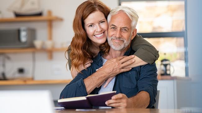 Portrait of happy mature couple at home looking at camera. Romantic wife embracing senior husband from behind while laughing together. Portrait of beautiful woman in love hugging  old man in perfect harmony. Retirement generic seniors