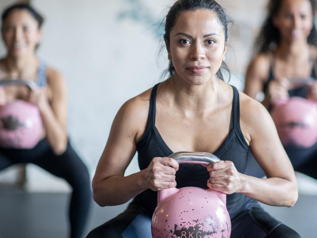 A multi-ethnic group of people are squatting while holding kettle weights.
