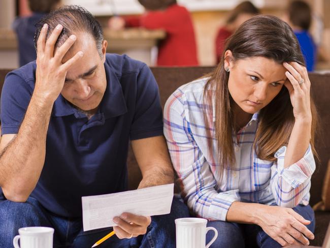 Latin descent man and woman struggle to pay their monthly bills.  They are calculating expenses versus budget income and are upset.  Many invoices, laptop on living room coffee table.  Children in kitchen background.  Mid-adult age couple.  Frustration among middle-class people.   Great imagery for election season:  home finances, recession, past due bills, mortgage, debt, stress, worry, taxes.