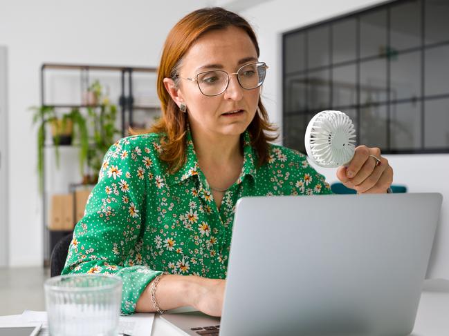 Tired mature businesswoman using laptop while sitting with portable electric fan at office desk