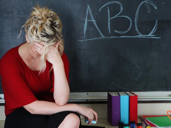 Elementary classroom setting with tired or frustrated teacher holding her head. She's sitting in front of an chalkboard with ABC