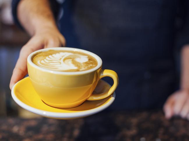 A barista serves up a hot espresso latte in a classy ceramic mug with heart shaped fern poured into the steamed milk foam. Vertical. Extremely shallow depth of field with selective focus on barista's finger.