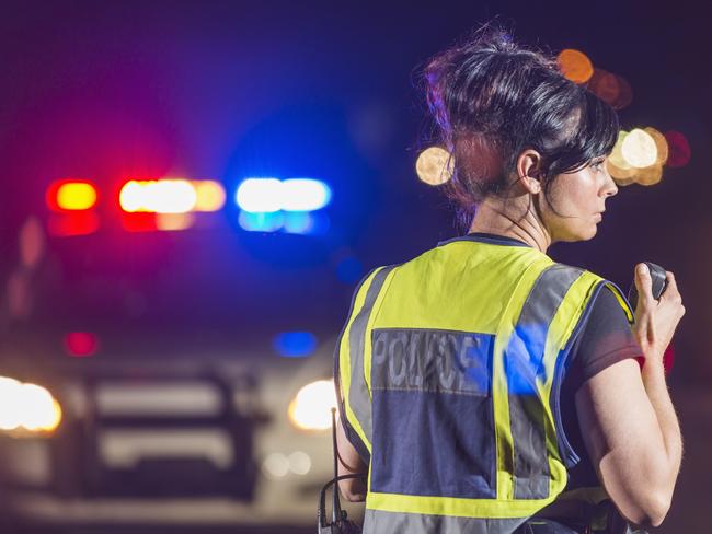 Rear view of a female police officer standing in the street at night, talking into her radio. Her patrol car is in the background with the emergency lights illuminated. She is wearing a yellow safety vest. Tablet generic