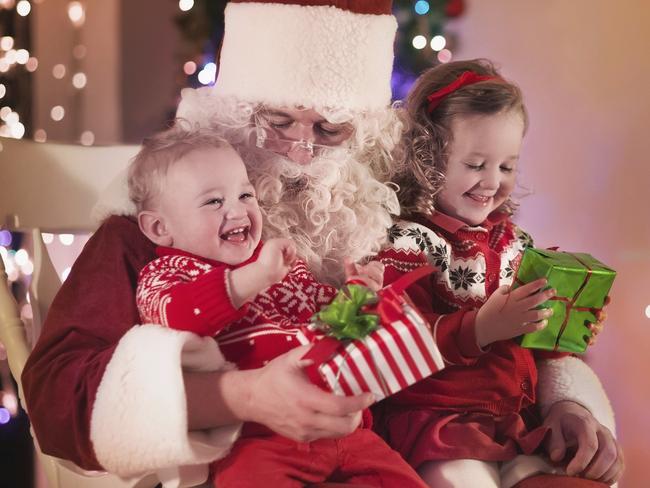 Santa Claus and children opening presents at fireplace. Kids and father in Santa costume and beard open Christmas gifts. Little girl helping with present sack. Family under Xmas tree at fire place.