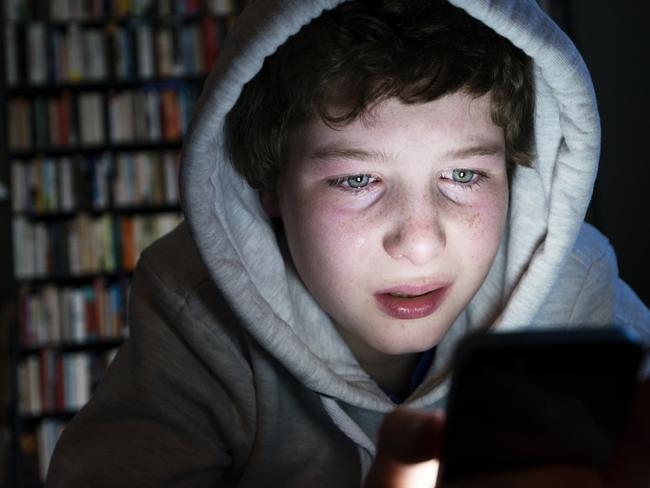 Candid colour portrait of a young teen in tears staring intently into a smart phone. His face is illuminated by the light from the digital device that he is holding, he is wearing a grey hoodie with the hood up and he has a down beat look on his face, maybe he is being bullied online or looking at something more sinister online. Horizontal format with a dark background that could be suitable for some copy space.