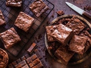 Top view of homemade chocolate brownies shot on dark table. A wooden plate filled with chocolate brownies is at the right and a cooling rack with brownies is at the left. Some chocolate bars are at the bottom. A brown bowl with cocoa powder is at the bottom left corner of an horizontal frame. Predominant color is brown. Low key DSRL studio photo taken with Canon EOS 5D Mk II and Canon EF 100mm f/2.8L Macro IS USM.