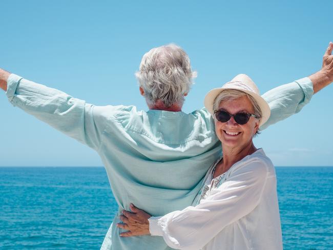 Cheerful senior woman with hat hugging her husband while looks at horizon over sea with outstretched arms. Happy retired couple enjoying summer holidays. Happy senior couple generic