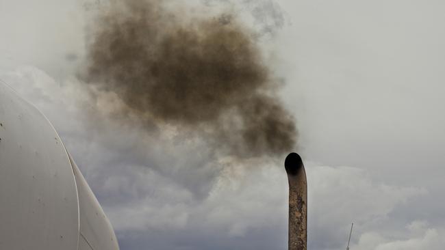 A white dump truck spews black exhaust smoke into a blue sky. Just the top of the truck is visible with the sky taking up half the image. The burning of fossil fuel by vehicles contributes to greenhouse gasses, pollution in urban city centers, and health concerns of people worldwide. Diesel fumes