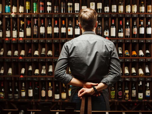 Bartender at wine cellar full of bottles with exquisite alcohol drinks that have various sweet and sour tastes and dates of manufacture on large wooden shelves.