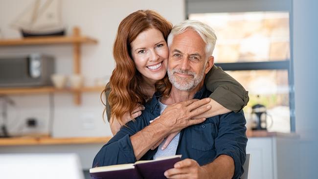 Portrait of happy mature couple at home looking at camera. Romantic wife embracing senior husband from behind while laughing together. Portrait of beautiful woman in love hugging  old man in perfect harmony. Retirement generic seniors