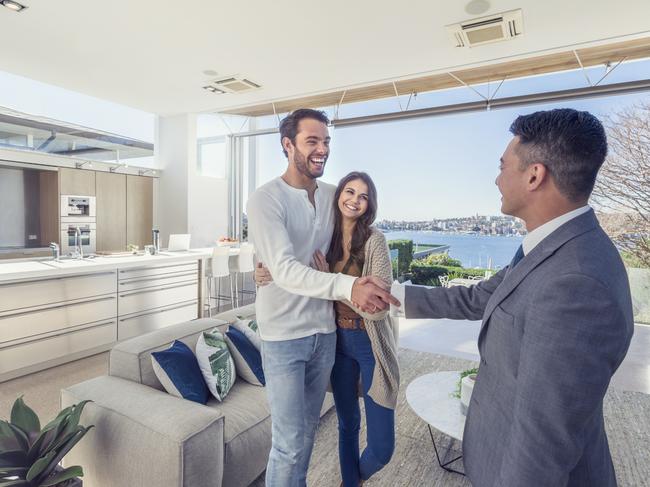 Real estate agent with couple in luxury home. They are shaking hands. There is a water view, kitchen and living room in the background. Couple are casually dressed. They are laughing. Agent is dressed in a suit and smiling. Wide angle. Picture: Istock