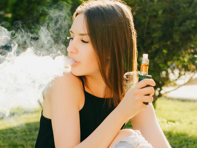 Outdoors photo of trendy attractive girl smoking vape, spending leisure time while sitting on grass in the park, with laptop.