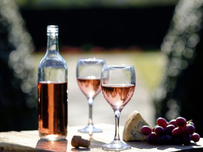 Rose wine filled bottle,glasses and a cork on a large cheese board with red grapes and stilton cheese with natural sunlight behind in a formal garden environment with copy space