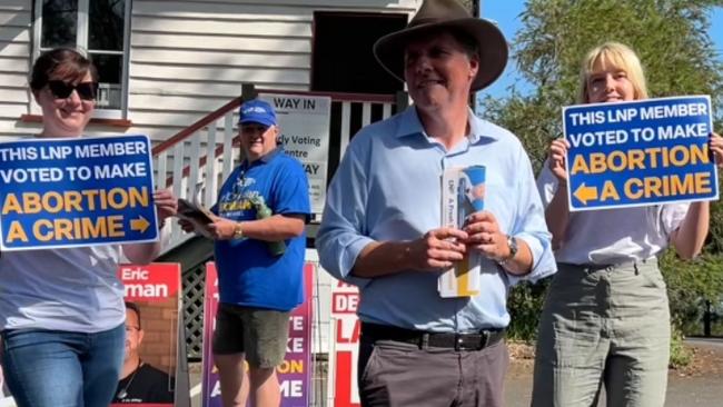 Moggill MP Christian Rowan flanked by protestors outside the electorate's pre-poll booth.