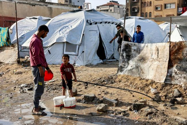 A youth speaks to a boy filling up buckets with water from a hose near tents at a shelter for displaced Palestinians in Gaza City