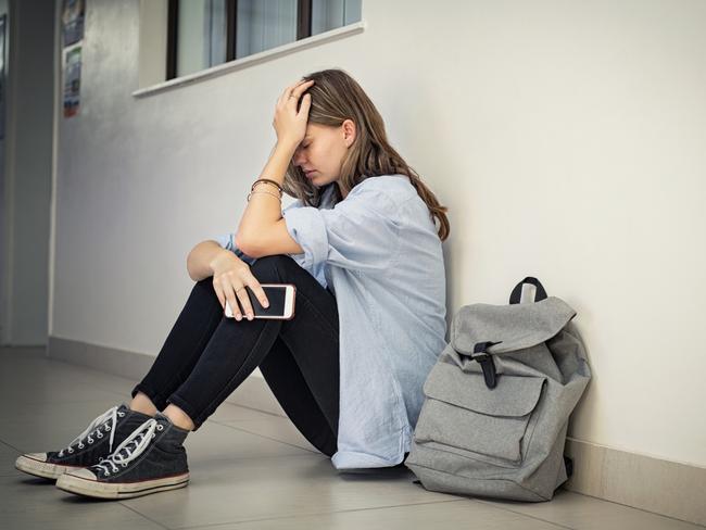 Upset and depressed girl holding smartphone sitting on college campus floor holding head. University sad student suffering from depression sitting on floor at high school. Lonely bullied teen in difficulty with copy space.