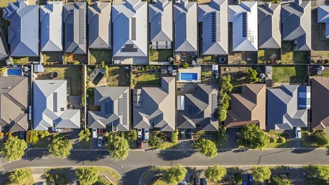 Aerial view directly above new outer suburban/semi-rural housing development with single-level housing between two streets with t-intersections and orange coloured street trees.  Mostly gray metal roofing, landscaped front and back yards, some solar panels, cars and motor vehicles parked in driveways and street, some street and parkland trees, water tanks in backyards, sheds and garages, swimming pools.  Mount Barker, South Australia; property investment housing money generic