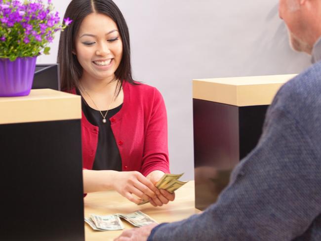 An adult man bank customer making a financial transaction with a bank teller over the counter in a retail bank. The woman Asian bank teller is smiling and cheerfully providing the customer service. Photographed behind the shoulder of the customer in a horizontal format.