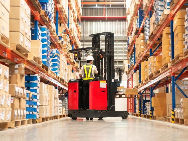 Worker in forklift-truck loading packed goods in huge distribution warehouse with high shelves.