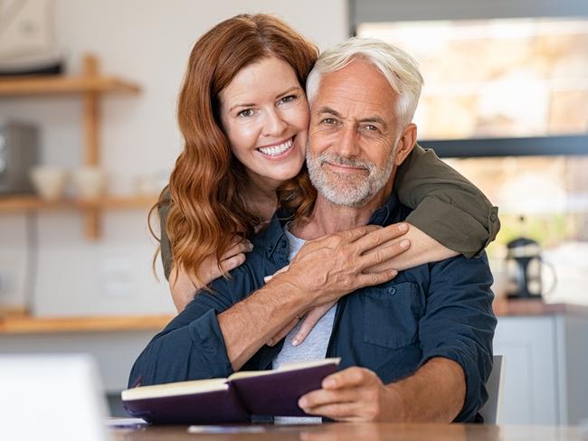 Portrait of happy mature couple at home looking at camera. Romantic wife embracing senior husband from behind while laughing together. Portrait of beautiful woman in love hugging  old man in perfect harmony. Retirement generic seniors