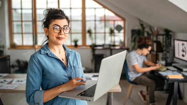 Portrait of young smiling woman in creative office, standing, holding laptop, looking at camera.