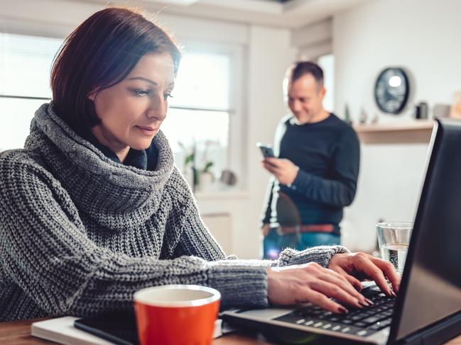 Woman wearing grey sweater using laptop at home office while her husband using smart phone in the background  working from home istock