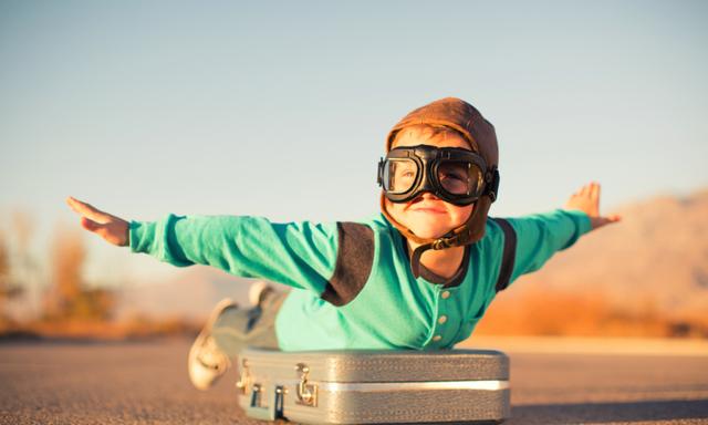 A young boy dressed in retro clothing and flying goggles dreams of flying on an exotic vacation at a far off destination. He is outstretching his arms like an airplane while on top of a suitcase and he has a happy expression on his face. Image taken in Utah, USA.