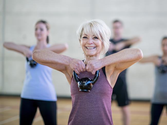 An older Caucasian woman is seen doing a squat with a kettlebell, while participating in a co-ed, multi-ethnic, fitness class. She is expressionless and focusing on her form. Active seniors generic