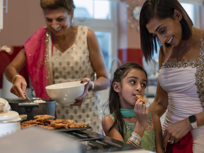 A close-up side view of a three gen female family cooking food for their family as they celebrate Diwali in the family home. The young girl is eating some fresh Jalebi from the baking tray and trying it as her grandmother serves the fresh food into serving dishes.