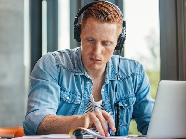 Portrait of young student wearing headphones sitting at the table in library and reading book. University student finding information for his academic assignment.