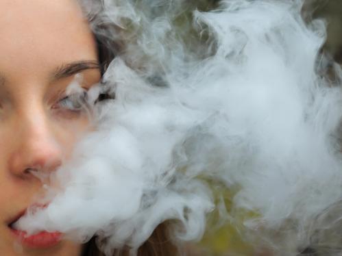 Vape teenager. Young pretty white girl in white cardigan smoking an electronic cigarette opposite destroyed brick wall on the street in the spring. Bad habit that is harmful to health.