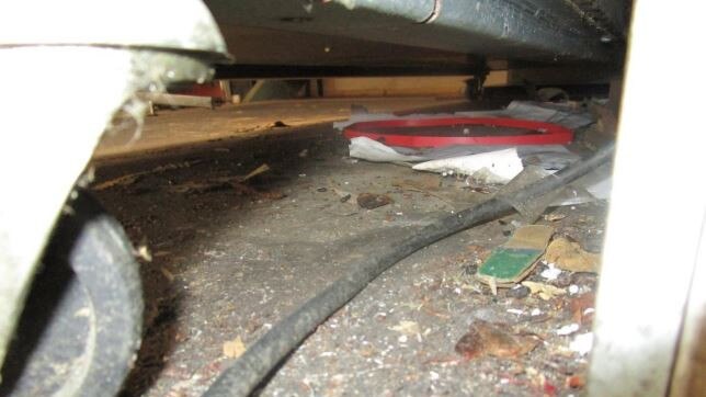 Unidentified waste cardboard, rodent droppings, and other unidentifiable visible matter on the floor underneath a chest freezer.