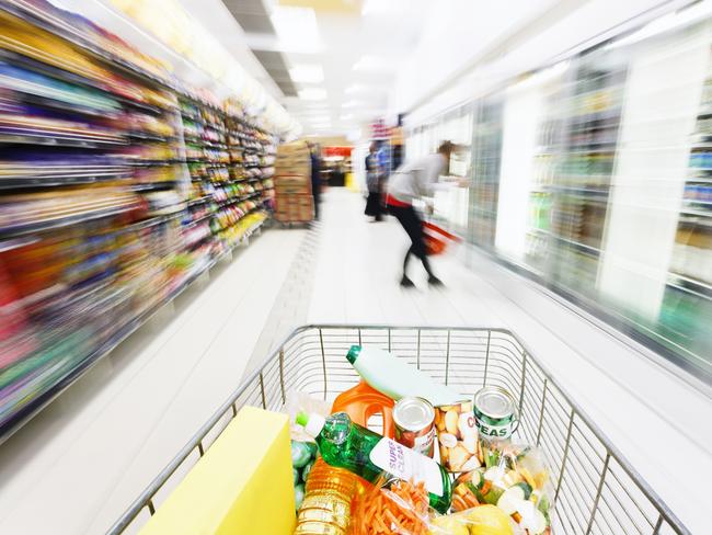View over filled shopping cart racing down a supermarket aisle towards out-of-focus  shoppers, showing extreme, multicolored motion blur.  The product labels in the cart are made by the photographer