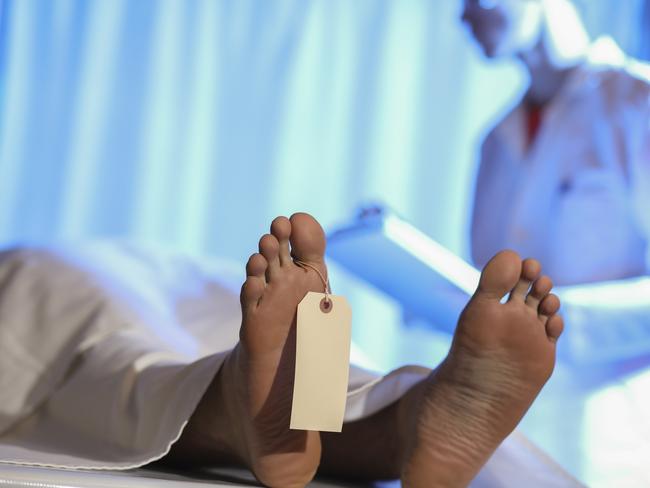 Medical examiner or forensic scientist with dead man's corpse in morgue.  He takes notes on medical chart after autopsy.Generic post mortem autopsy photos - iSTOCK