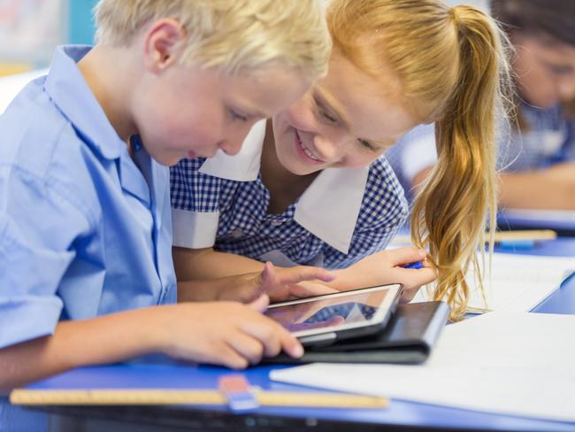 Boy and girl students sharing a digital tablet. They are in a classroom wearing school uniforms. Both are excited, and one is smiling. Ethnic students in the background.