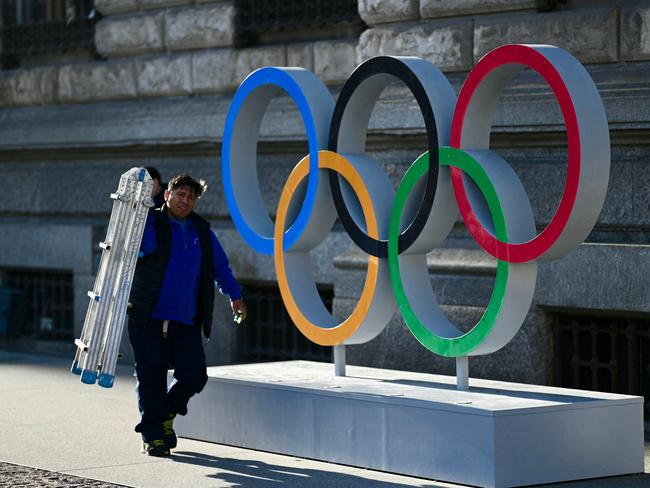 A man carrying a lader walks past the Olympic rings set at the entrance of Milan's city all on February 14, 2024 in Milan. Milan and Cortina will host the 2026 Winter Olympic Games. (Photo by GABRIEL BOUYS / AFP)