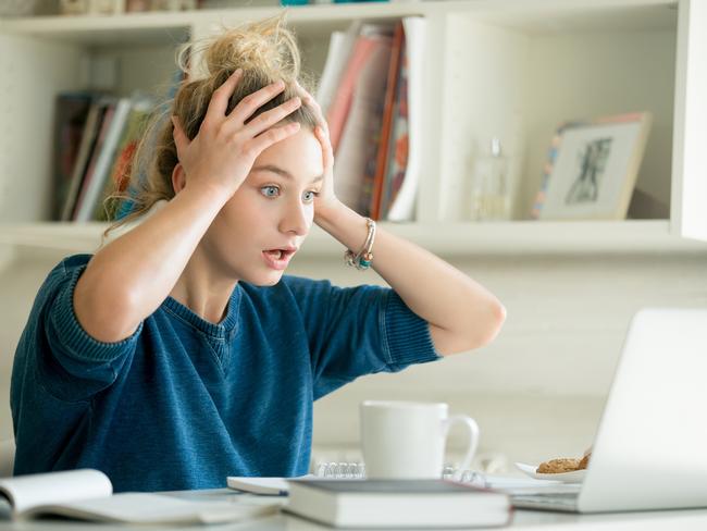 money mistakes - generic woman shocked, stressed... Portrait of an attractive woman at the table with cup and laptop, book, notebook on it, grabbing her head. Bookshelf at the background, concept photo