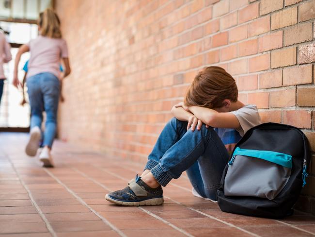 Little boy sitting alone on floor after suffering an act of bullying while children run in the background. Sad young schoolboy sitting on corridor with hands on knees and head between his legs.