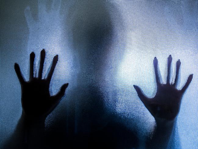 Colour backlit image of the silhouette of a woman with her hands pressed against a glass window. The silhouette is distorted, and the arms elongated, giving an alien-like quality. The image is sinister and foreboding, with an element of horror. It is as if the 'woman' is trying to escape from behind the glass. Horizontal image with copy space.