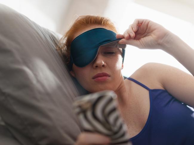 Young woman with sleep mask waking up in bed and snoozing alarm clock on cell phone, mobile telephone. Girl sleeping in bedroom at home and waking up after receiving text message or call on smartphone