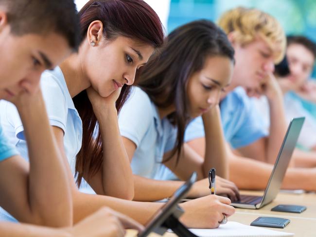 Row of private high school students work on assignment in class. They are writing or using laptops or digital tablets. They are concentrating as they study. They are wearing school uniforms. Photo: istock