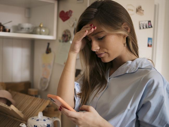 Young caucasian woman holding her forehead while scrolling through social media on her smart phone. Female looking worried using mobile phone in kitchen at home.   istock image