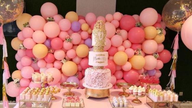 The tiara-topped cake surrounded by various desserts in front of the balloon wall.