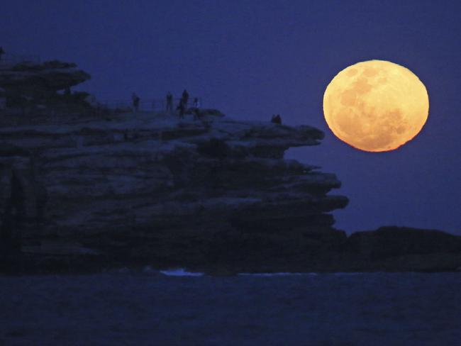 The moon rises with an orange glow as people watch from a rock cliff at Bondi Beach on Tuesday. AP Photo/Rick Rycroft